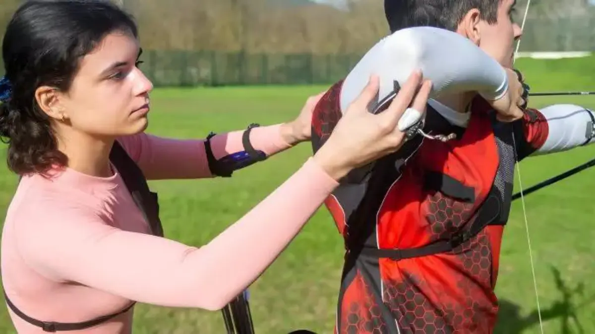woman wearing pink top assists another archer which his shoulder positioning while getting ready to shoot his arrow