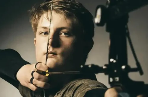 young light haired man steadily holds his bow draw weight and arrow while aiming ahead