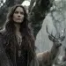 Celtic goddess with flowing dark brown hair standing deep within an ancient forest, with a stag stands nearby